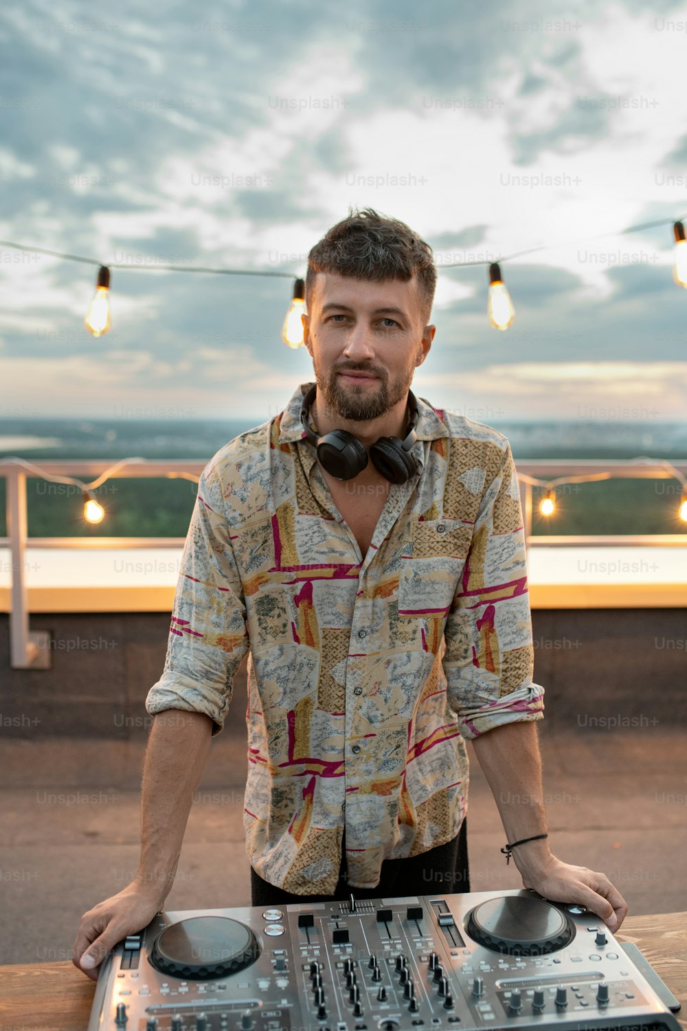 Young bearded deejay standing by soundboard in front of camera at rooftop patio