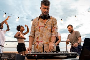 Joven DJ con auriculares alrededor del cuello de pie junto a la caja de resonancia frente a amigos bailando