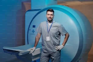 Clínico confiante em uniforme cinza em pé por mesa de equipamentos de varredura de ressonância magnética em laboratório moderno