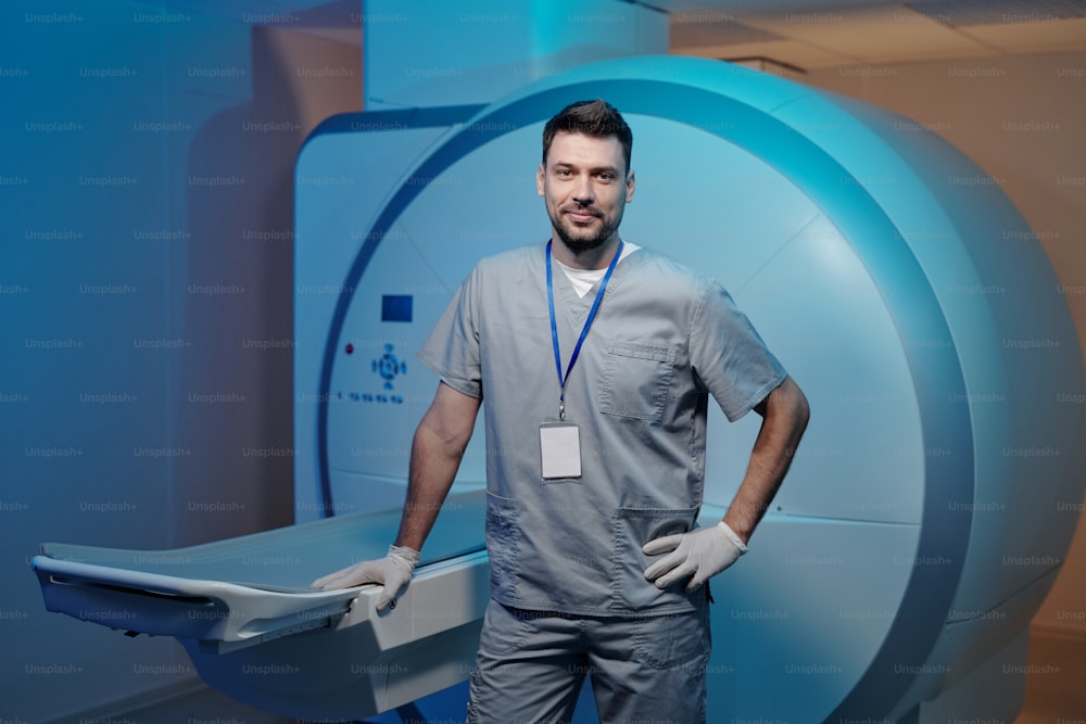 Medico fiducioso in uniforme grigia in piedi vicino al tavolo dell'apparecchiatura di scansione MRI in un laboratorio moderno