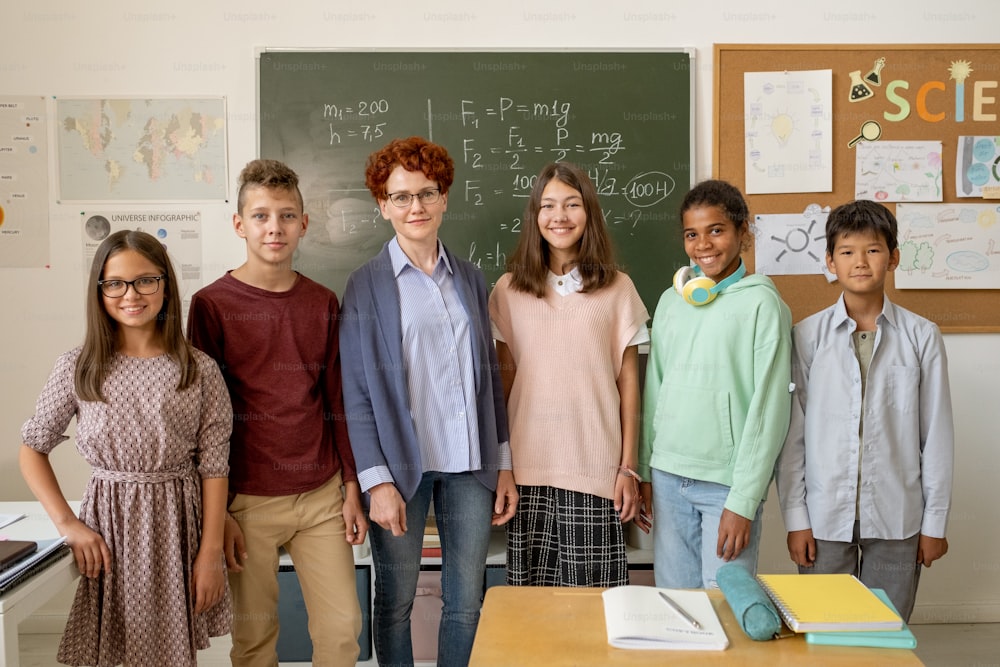 Row of happy young teacher and clever schoolchildren standing by blackboard in classroom