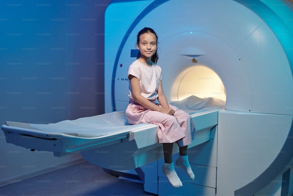 Piccolo paziente sorridente seduto sul lungo tavolo medico prima o dopo la procedura di risonanza magnetica