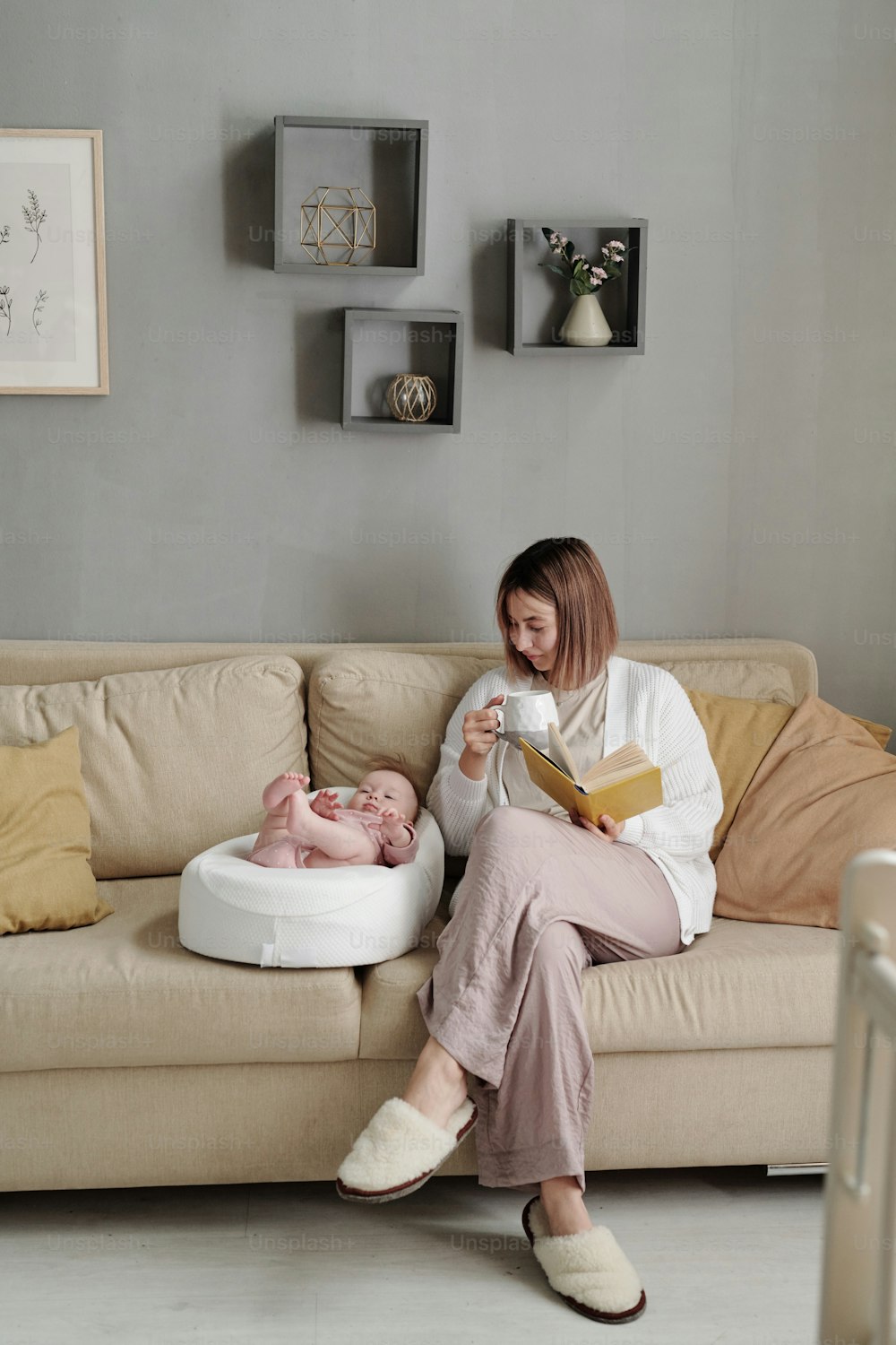 Young woman with cup of tea and book sitting on couch by her baby daughter sleeping in small soft bed