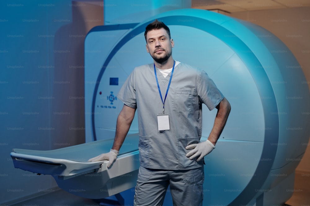 Giovane radiologo guantato contemporaneo in uniforme grigia in piedi vicino alle apparecchiature mediche
