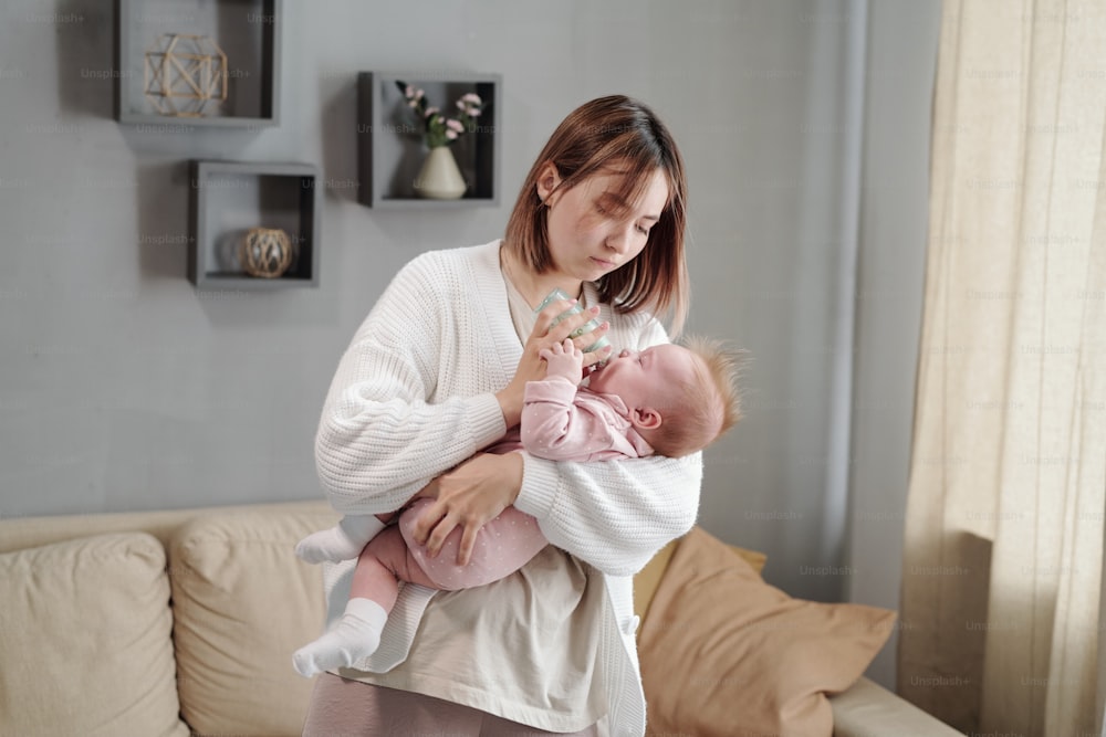 Young woman holding small bottle while feeding her baby daughter in home environment