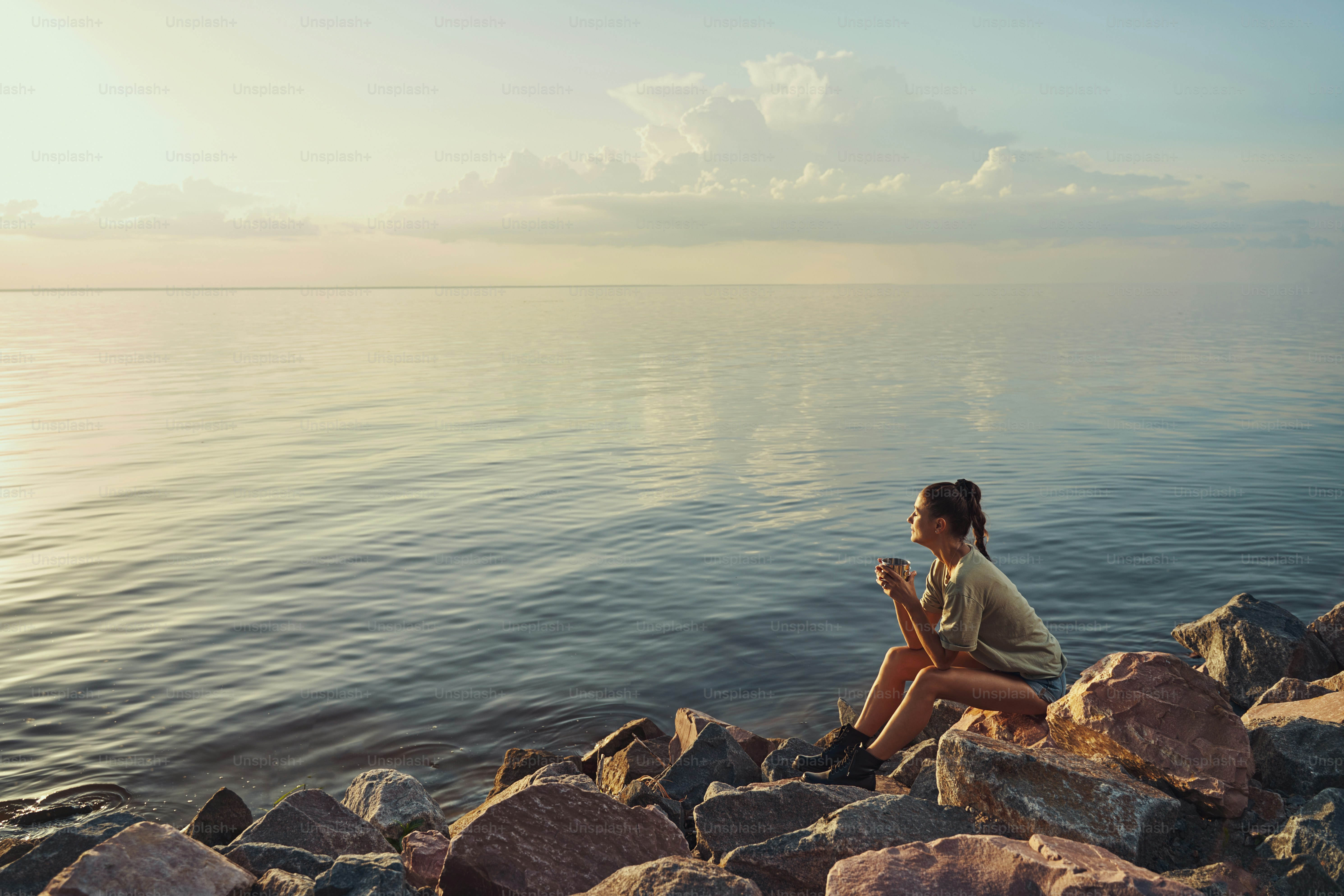 Woman resting on big stone at lake coast with mug of tea in her hands while enjoying sunset