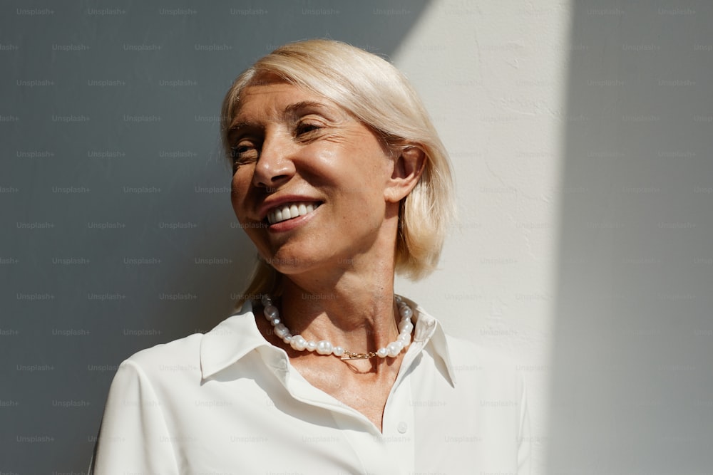 Minimal portrait of elegant mature woman lit by sunlight against white wall and smiling, copy space