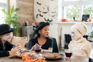 Zwei Mädchen und Jungen in Halloween-Kostümen, die Getränke mit Keksen trinken, während sie am Tisch interagieren