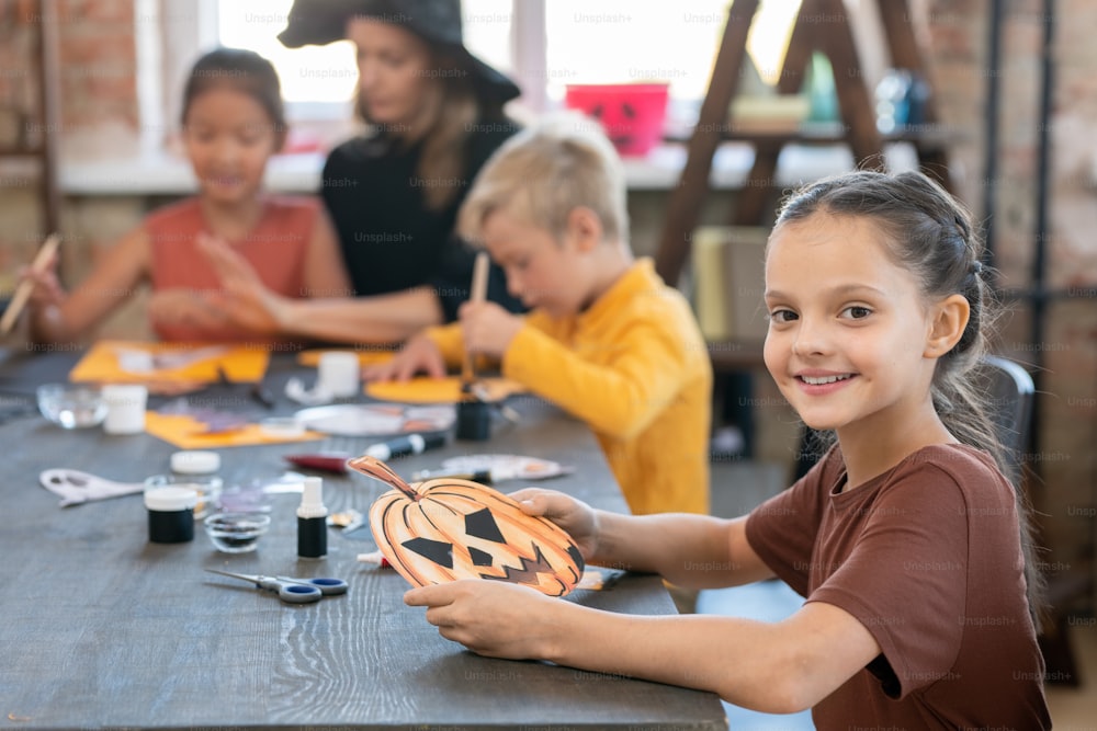 Menina feliz com símbolo de halloween sentado à mesa contra professor e colegas de classe