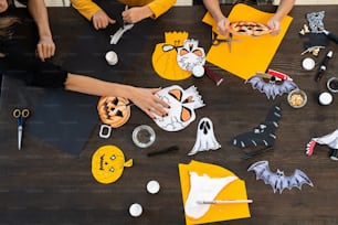 Hände von Kindern und jungen Frauen, die Halloween-Papiersymbole zusammen am Tisch malen