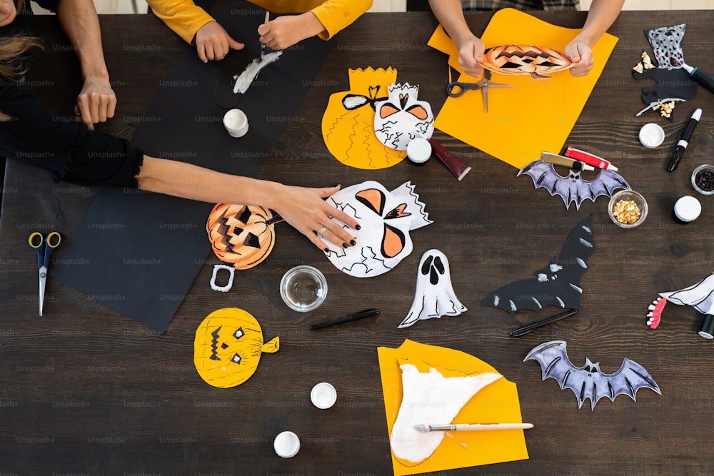 Hände von Kindern und jungen Frauen, die Halloween-Papiersymbole zusammen am Tisch malen