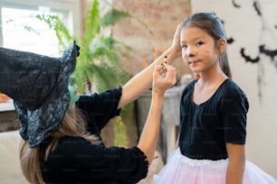 カメラの前でかわいいアジアの女の子の顔にハロウィーンの化粧をする魔女の帽子と黒いプルオーバーを着た若い女性