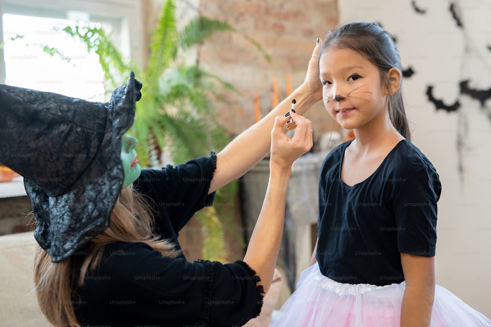 Young female in witch hat and black pullover applying halloween makeup on face of cute Asian girl in front of camera