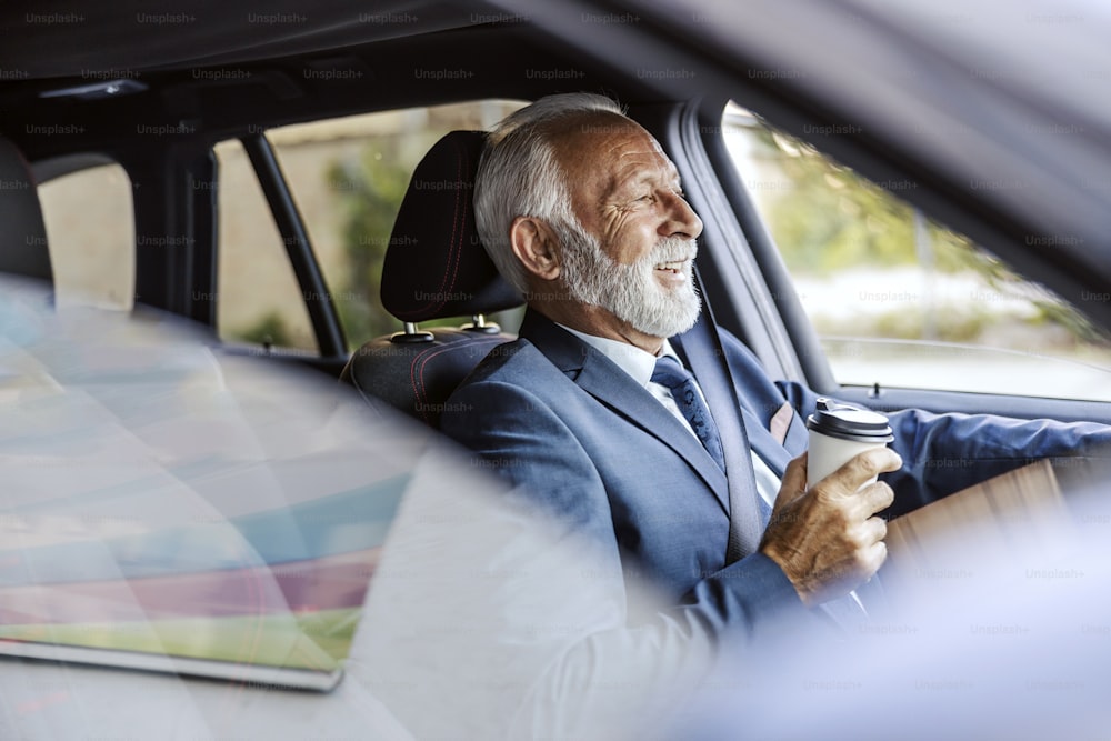 Un vecchio uomo d'affari barbuto in giacca e cravatta sta guidando la sua auto per andare al lavoro e bevendo il suo caffè mattutino per andare.