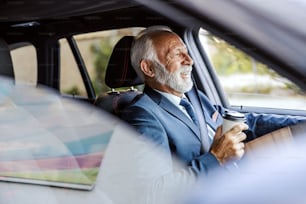 Un viejo hombre de negocios barbudo con traje conduce su automóvil al trabajo y bebe su café de la mañana para llevar.