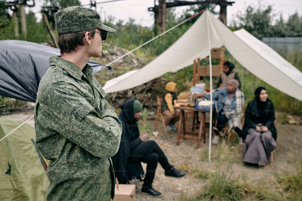Joven soldado serio con uniforme militar y gafas de sol de pie con los brazos cruzados y mirando a los refugiados mientras los salvaguardan en el campamento
