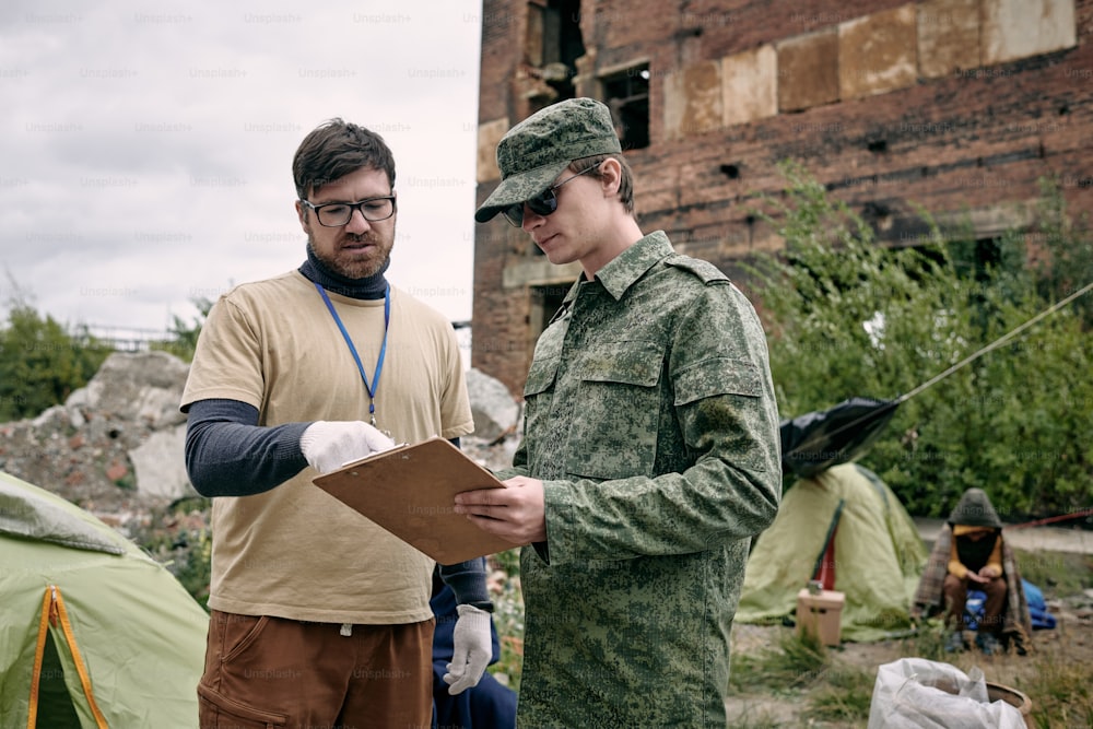 Trabajador social barbudo con guantes señalando la lista mientras habla con un soldado en el campo de refugiados
