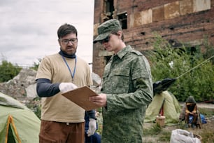 Bearded social worker in gloves pointing at list while talking to soldier in refugee camp