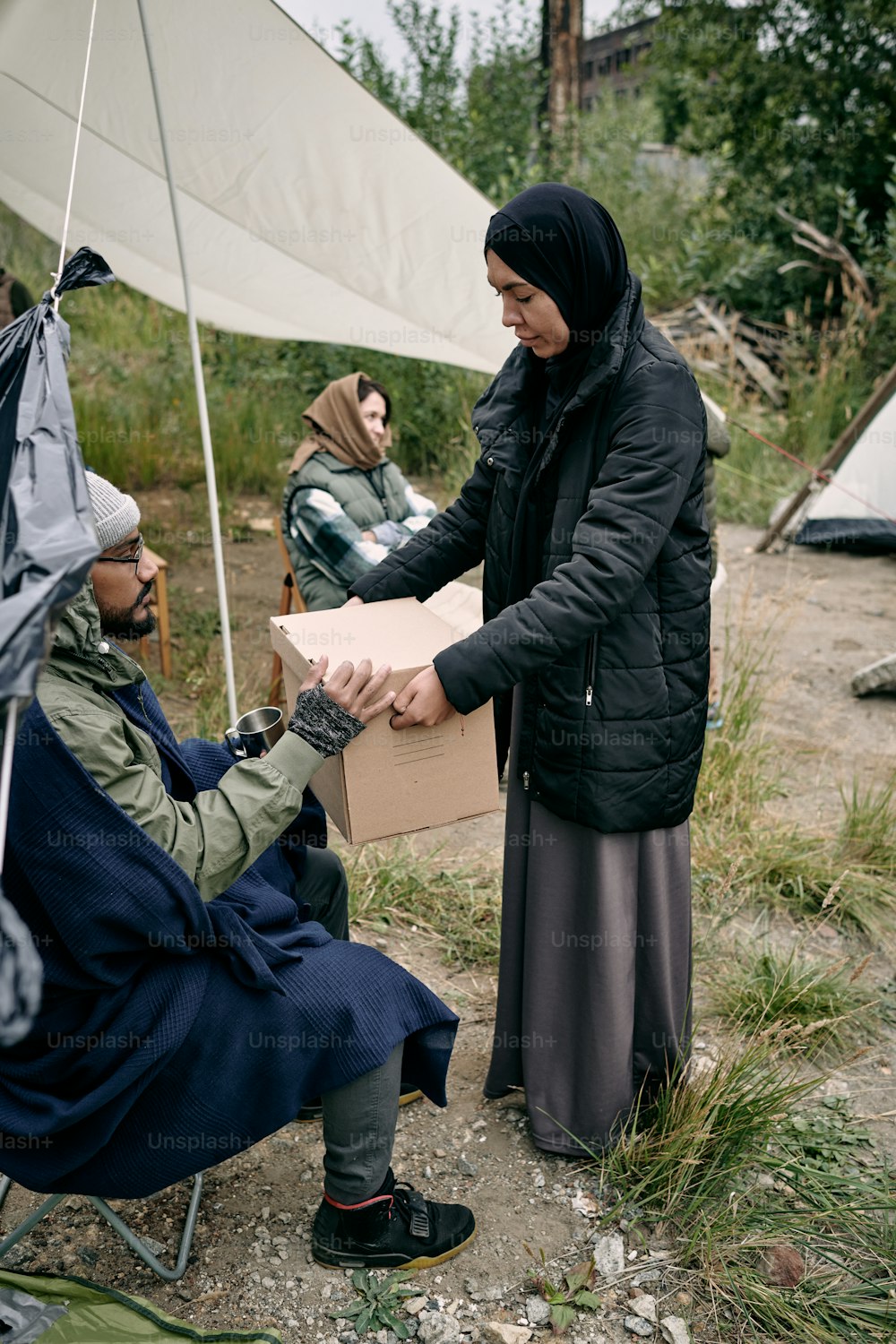 Muslim woman in black headscarf and big-size jacket sharing stuffs with young refugee in migrant camp