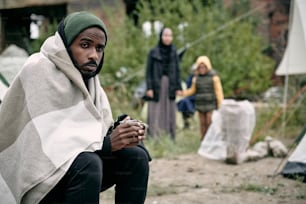 困惑した若い黒人男性が毛布に包まれ、移民キャンプに座って寒い季節にお茶を飲む