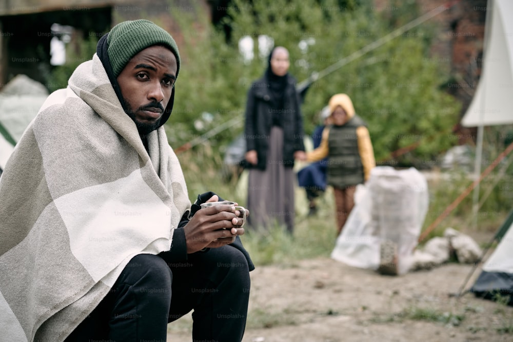 Puzzled young Black man wrapped into blanket sitting in migrant camp and drinking tea in cold weather