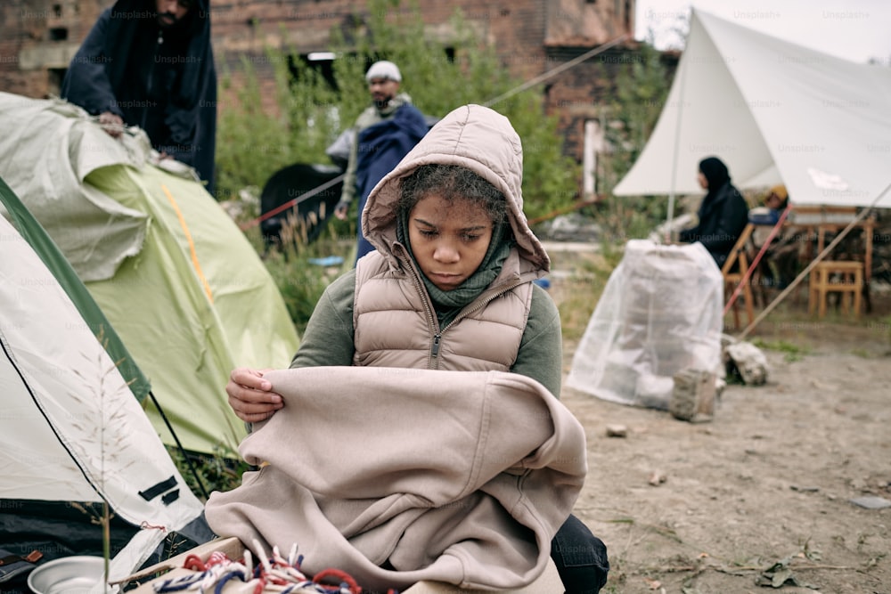 Sad middle-eastern refugee girl in hooded vest holding plaid while freezing in tent camp for migrants