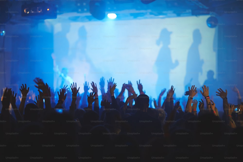 Rear view of audience going crazy at concert, hands up in the air in dark blue-lit music hall