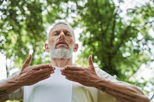 Low angle view of the elderly man keeping his eyes closed and doing breathing exercise while enjoying of the nature and meditating. Sport concept