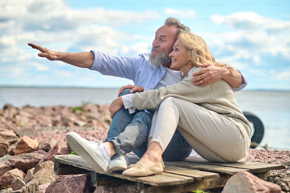 Curious, interesting. Delighted smiling bearded man hugging blonde woman pointing with hand forward sitting on seashore on fine warm day