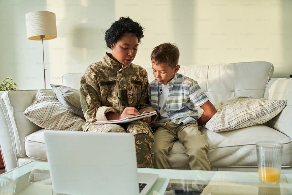 Black mother writing in notebook and showing it to her son. Concept of motherhood and family relationship. Female soldier wearing camouflage uniform. People sitting on couch in living room at home