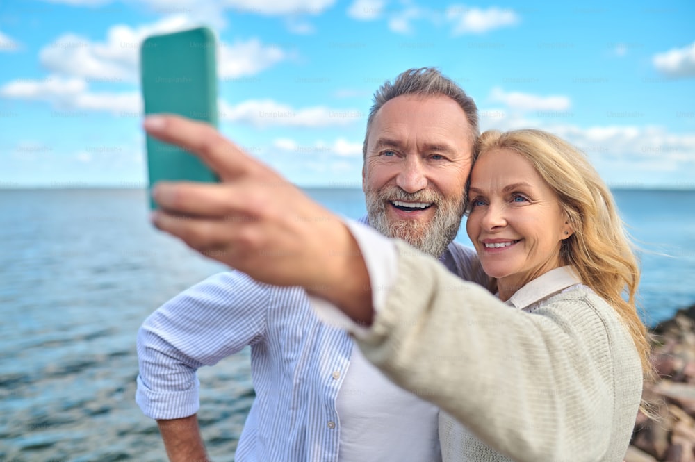 Buena foto. Mujer bonita sonriente con teléfono inteligente en la mano extendida y hombre barbudo alegre tomándose selfie en la naturaleza cerca del mar