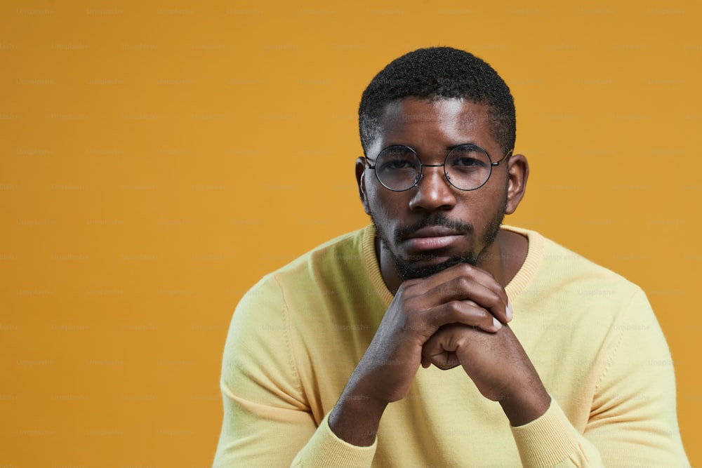 Retrato mínimo de homem afro-americano usando óculos e olhando para a câmera enquanto descansa queixo na mão e posando contra fundo amarelo, espaço de cópia