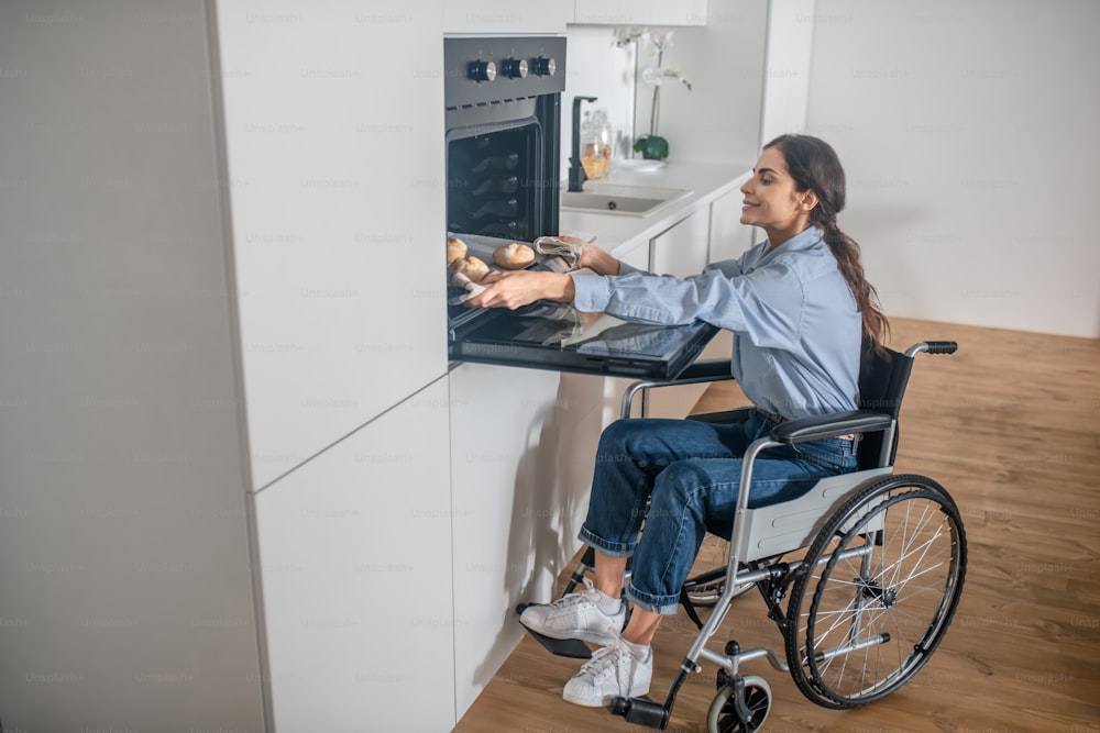 Cooking. A girl on a wheelchair opening oven in the kitchen while cooking soemthing