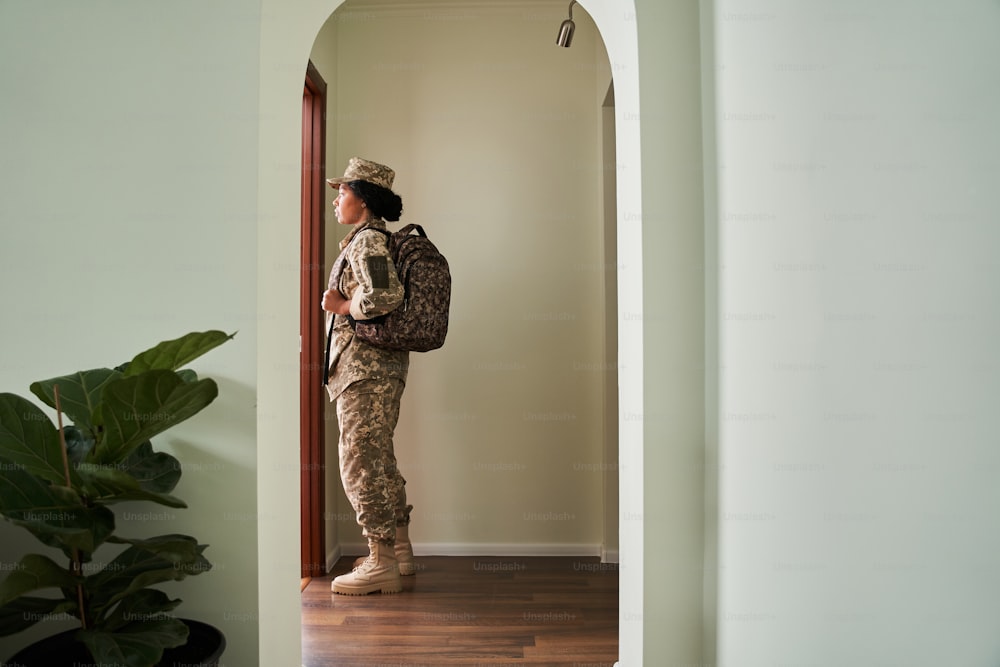 In den Krieg. Ganzkörperansicht der Soldatin, die eine Militäruniform trägt, einen Rucksack hält und mit traurigen Emotionen auf die Tür schaut, während sie zum Kampf geht