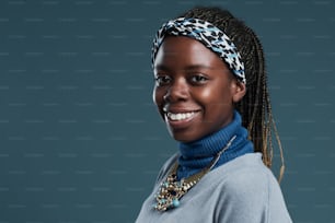 Retrato mínimo de una mujer afroamericana sonriente con pañuelo en la cabeza y joyas afro mientras está de pie contra un fondo azul profundo, espacio de copia