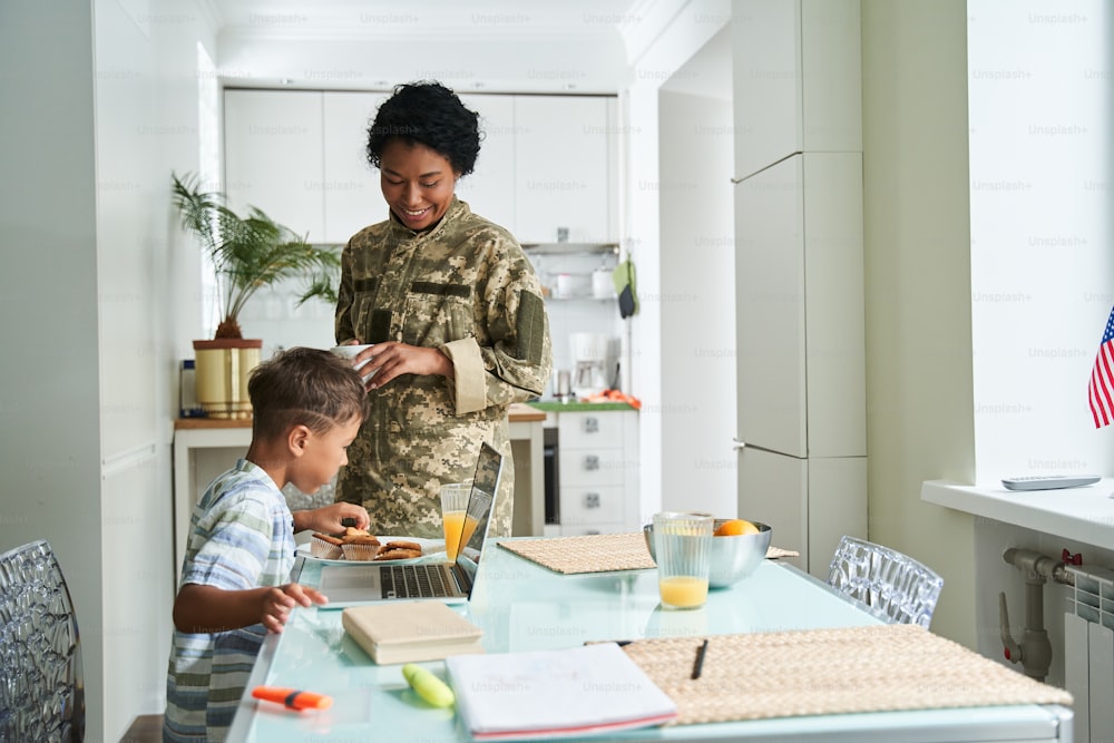 Così affamato. Donna soldato in uniforme militare che guarda il suo figlioletto mentre è seduto al tavolo e mangia biscotti per la sua colazione