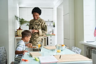 So hungry. Soldier woman in military uniform looking at her little son while he sitting at the table and eating cookies for his breakfast