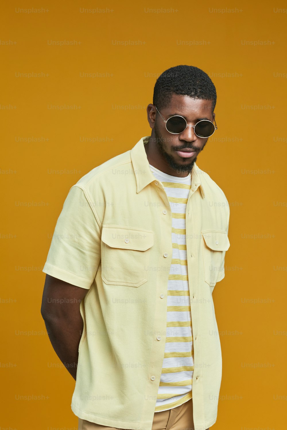 Retrato vertical da cintura para cima do homem afro-americano bonito usando óculos escuros enquanto posa contra o fundo amarelo no estúdio