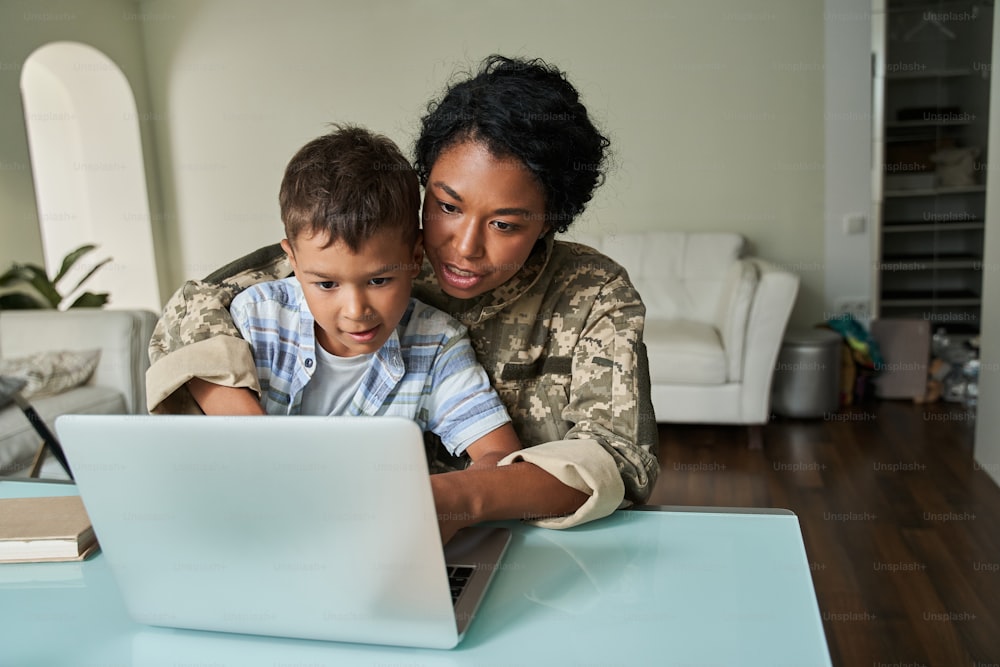 So interesting. Waist up portrait view of the little boy sitting at the knees of his military mother and looking at the laptop screen together. Family spending weekend at home