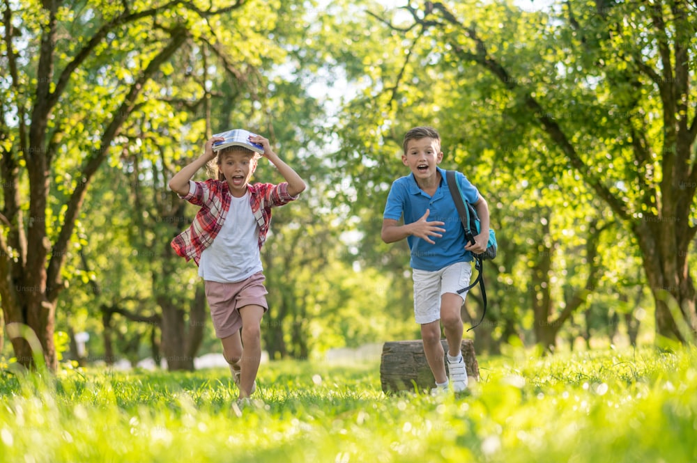 Colpo di scena a sorpresa. Ragazzo emotivo allegro con lo zaino e la ragazza con il libro sopra la testa che corre con la bocca aperta nel parco verde in una giornata di sole