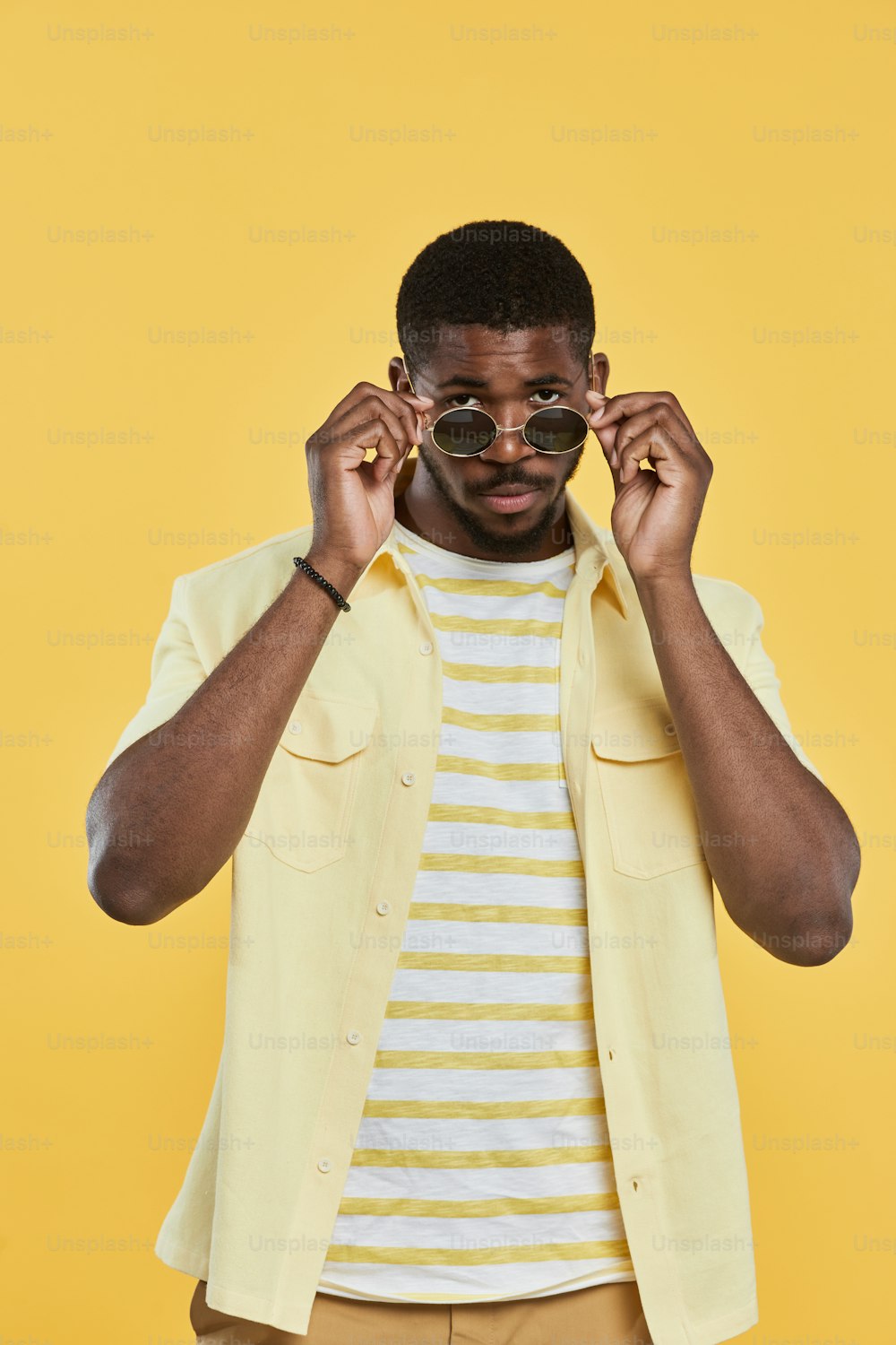 Vertical portrait of trendy African-American man wearing sunglasses and looking at camera while standing against yellow background in studio