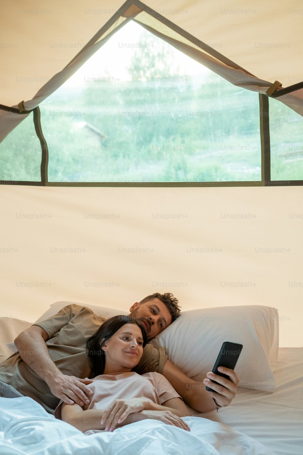 Young restful couple with smartphone making video call while relaxing in bed inside glamping house