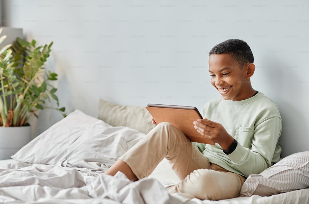 Full length portrait of smiling African-American teenager using tablet while sitting on bed in cozy room, copy space