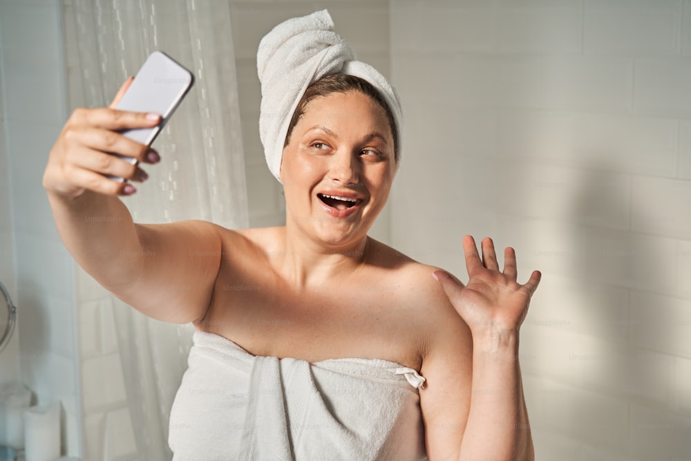 Mulher oversize tirando selfie e acenando com a mão no celular. Conceito de cuidados com a pele corporal e higiene. Menina jovem europeia sorridente com toalha enrolada na cabeça. Interior do banheiro em apartamento moderno