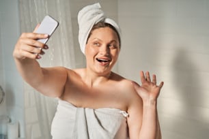 Mujer de gran tamaño tomando selfie y agitando la mano en el teléfono móvil. Concepto de cuidado e higiene de la piel corporal. Joven europea sonriente con toalla envuelta en la cabeza. Interior del baño en apartamento moderno