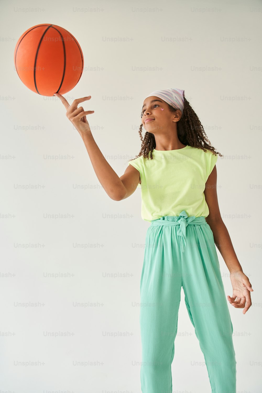 Black little girl spinning basketball ball on her finger. Smiling brunette schoolgirl with heart shaped sticker on her face. Concept of childhood. Isolated on grey background. Studio shoot. Copy space