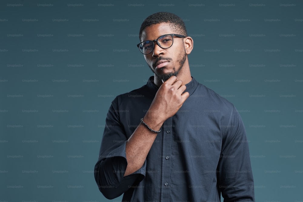 Waist up portrait of confident African-American man wearing glasses looking at camera while standing against deep blue background, copy space