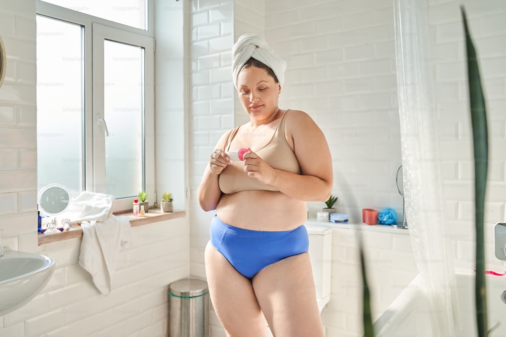 Vista de cuerpo entero de la mujer con lencería mirando su antitranspirante mientras está parada en el baño. Foto de archivo