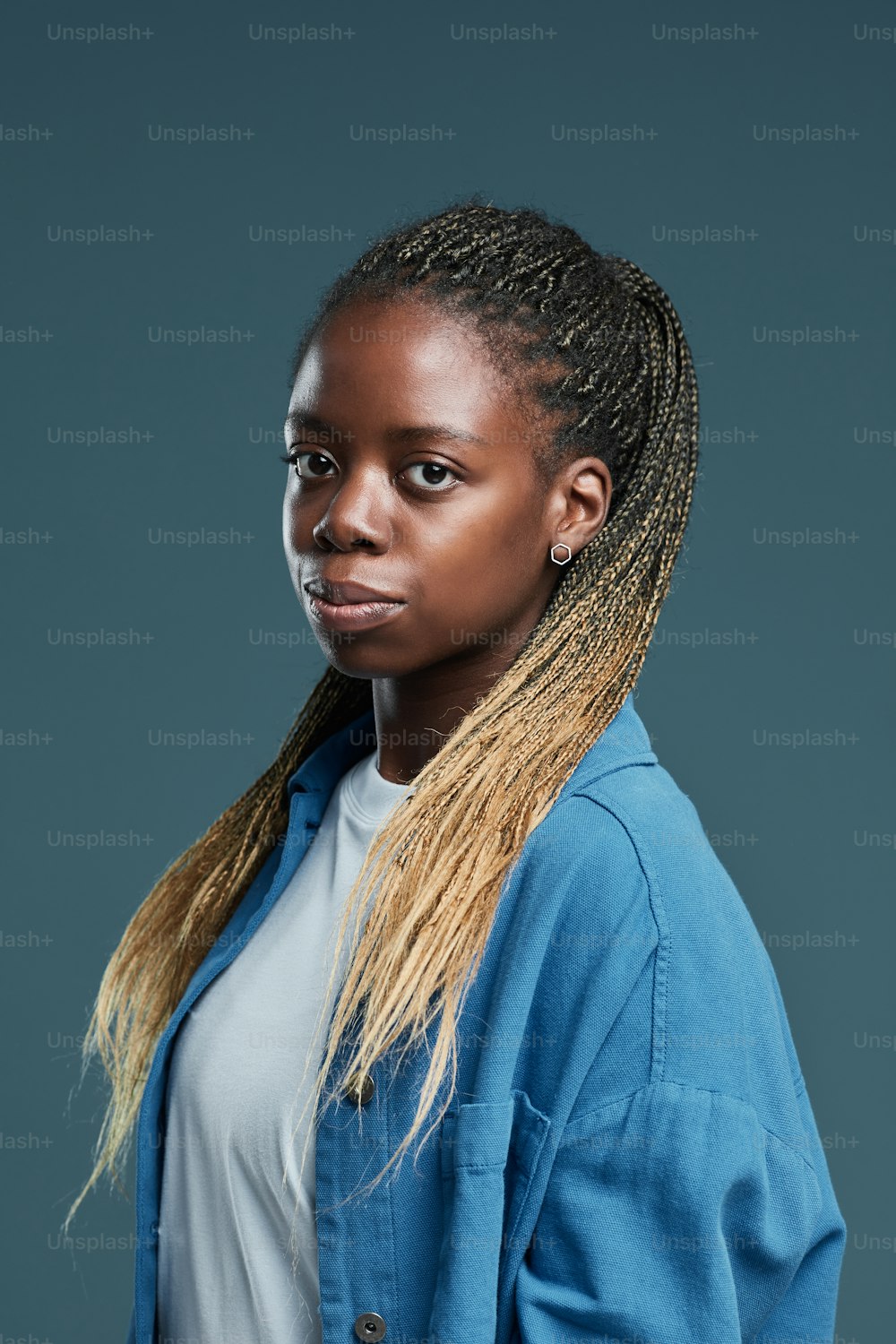 Retrato vertical de una joven afroamericana moderna con peinado afro trenzado mirando a la cámara mientras posa contra fondo azul