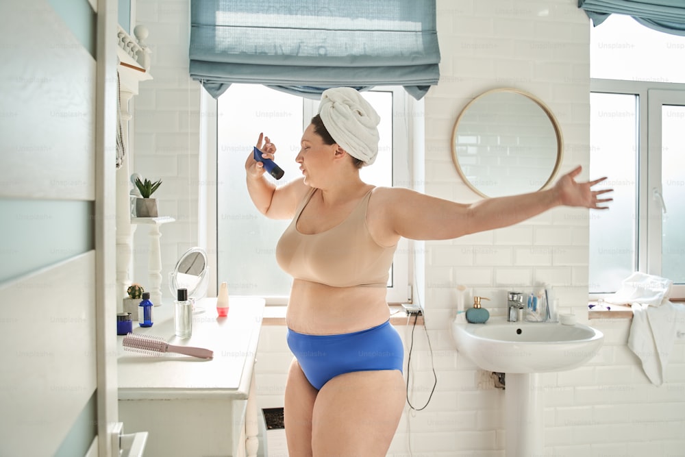Oversize woman singing in tube with cosmetic cream like in microphone and dancing. Concept of body skin care and hygiene. Young european girl with wrapped towel on head. Bathroom interior